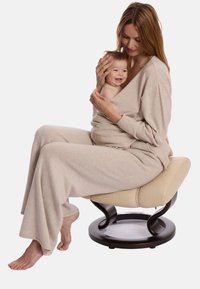 Un ensemble de loungewear en tricot beige présente un décolleté en V profond et des manches longues, porté par une femme tenant un bébé. La scène comprend un tabouret en bois courbé.