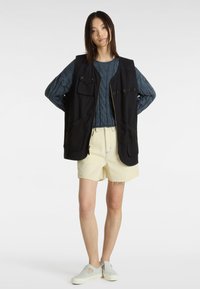 Gilet noir avec plusieurs poches, porté sur un pull en maille bleue texturé, assorti à un short jaune clair effiloché et des chaussures gris à enfiler.