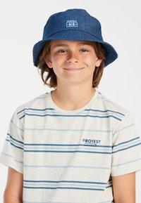 Blauwe denim bucket hat met een brede rand, voorzien van een logo patch aan de voorkant. Het model draagt een beige gestreepte T-shirt met blauwe lijnen.