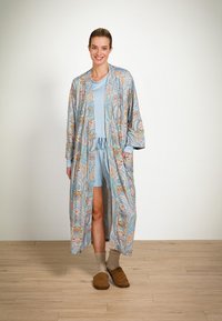 Lichtblauwe kimono-achtige badjas met bloemmotieven, gedragen over een bijpassende korte set. Zachte textuur, lange mouwen en zijzakken. Bruine slippers.