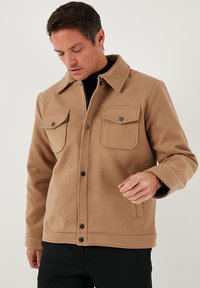 Beżowa kurtka z mieszanki wełny, z kołnierzem, dwoma kieszeniami na piersi, zatrzaskami i prostym krojem. Tkanina wydaje się miękka i teksturowana.