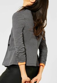 Černo-bílý vzorovaný blazer s vypasovaným střihem, nařasenými rukávy a oranžovými akcenty na manžetách, spárovaný s černými kalhotami.