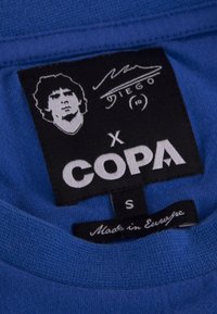 Blauwe t-shirt label met zwart textiel etiket waarop het portret van Diego Maradona staat, witte tekst voor het merk "COPA," maat "S," en "Gemaakt in Europa."