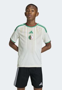 Maillot de football blanc avec des accents verts et rouges, présentant des rayures verticales et l'emblème de l'Algérie. Associé à un short noir, avec trois rayures sur les côtés.