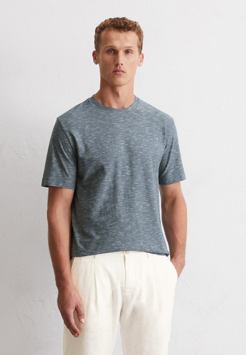 Kurzärmliges T-Shirt mit Rundhalsausschnitt aus strukturiertem blau-grauem Stoff, kombiniert mit hellen Leinenhosen, die eine entspannte Silhouette schaffen.