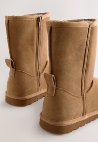 Barna szarvasbőr középhosszúságú csizma oldalán cipzárral, megerősített varrással és texturált gumitalppal; hátul húzófüllel ellátva.