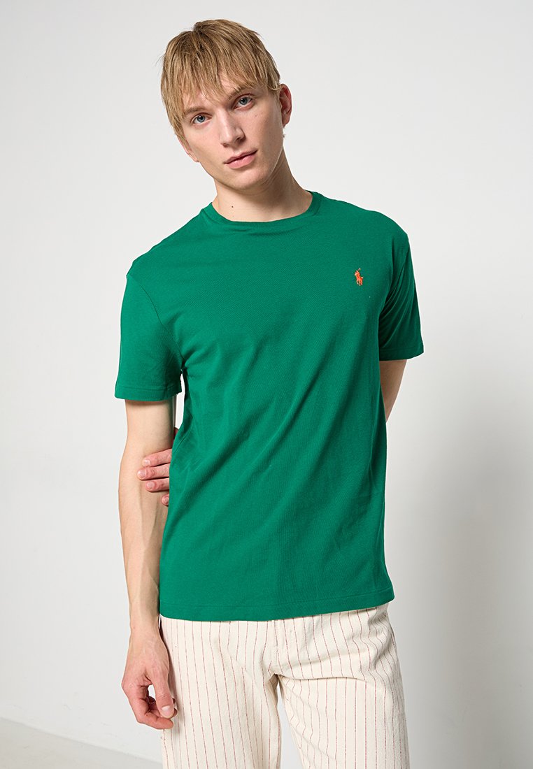 Zielona bawełniana koszulka z krótkim rękawem, okrągłym dekoltem i małym pomarańczowym logo na lewej piersi. Nosi się ją z beżowymi spodniami w paski.