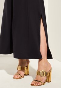 Svart maxiklänning med sidslitsar, matchad med sandaler med guldfärgad blockklack som har bred rem och spänne. Slät tygtextur.