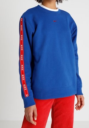 Persoon draagt een blauwe Nike-trui met rood logo en gebrande strepen op de mouwen, gecombineerd met een rode broek, tegen een effen achtergrond.