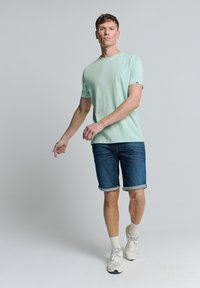 Lichtgroene T-shirt met korte mouwen, gecombineerd met op maat gemaakte denim shorts met omgeslagen mouwen en witte sportsneakers met grijze en gele accenten.
