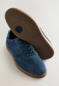 Sko i blåt ruskind med en glat tekstur, rundet tå, snøredesign og en brun gummisål. Logo prægning på sålen.