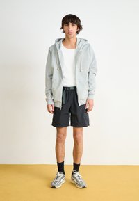 Pelēka kapuce ar auklītēm, balts bezpalaga topiņš, melnas šortas un pelēki skriešanas apavi. Ikdienas apģērbs, mīksta tekstūra un funkcionāli dizaina elementi.