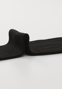 Chaussette noire en tricot avec une texture côtes, présentant un bord roulé et une finition lisse. Le matériau semble doux et flexible.