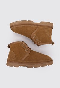 Botines de ante marrón con cordones en la parte delantera y suelas de goma gruesas, mostrados uno al lado del otro sobre un fondo gris.