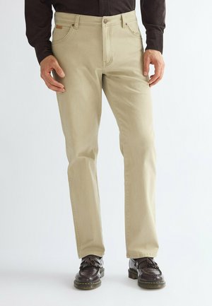 Man wearing beige straight-leg pants with front pockets and brown tassel loafers standing against a white background.