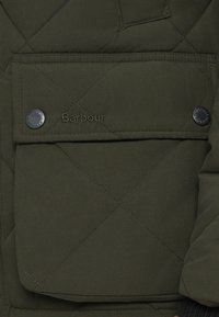 Casaco acolchoado verde-oliva com bolso de aba com botões prateados e logótipo "Barbour" em fio da mesma cor. Tecido texturado com costuras limpas.