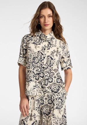 Kobieta ubrana w beżową koszulę z krótkim rękawem zapinaną na guziki oraz pasujące spodnie z czarnym kwiatowym i winoroślowym wzorem, stojąca z jedną ręką w kieszeni.