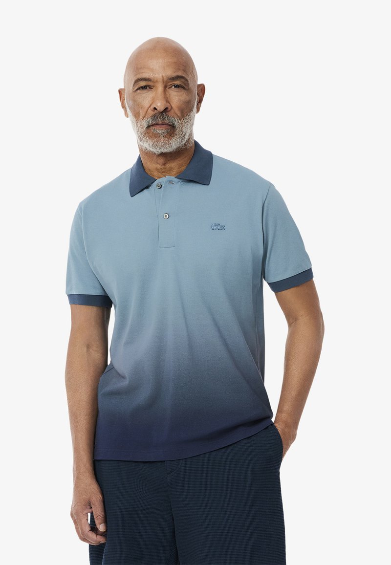 Pikétröja med en gradient från ljusblått till mörkblått, med en mörkblå krage, korta ärmar och en liten broderad logotyp på bröstet.