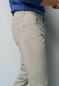 Beige katoenenbroek met een slim fit, frontzakken en een enkele achterzak met een bruine accentknop, weergegeven tegen een lichte achtergrond.