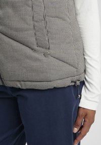 Kvadrični prsluk s crno-bijelim houndstooth uzorkom, s metalnim gumbima na kopčanju i podesivim vezicom na rubu.