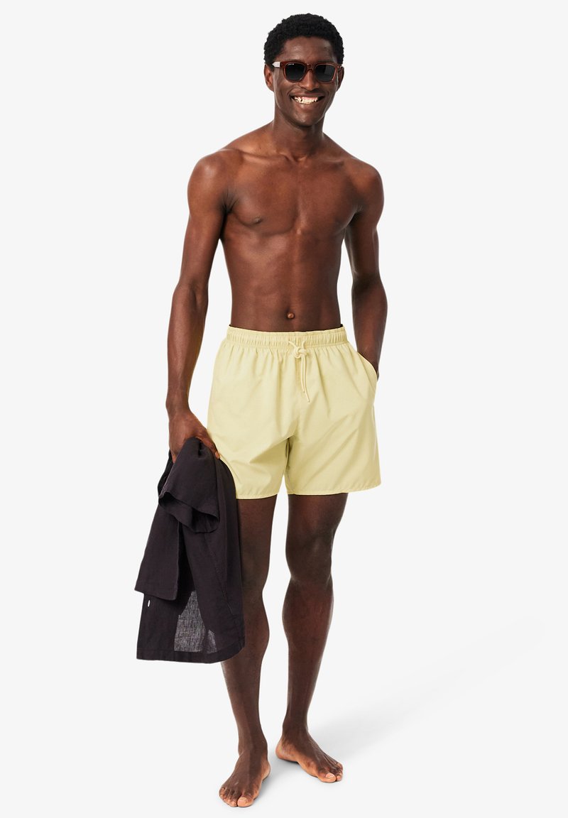 Glimlachende man zonder shirt, met zonnebril en lichtgele zwembroek, houdt een zwart shirt vast en staat op blote voeten tegen een witte achtergrond.