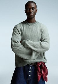 Hellgrüner, gerippter Pullover mit übergroßen Ärmeln, getragen über einem Jeansrock mit großen Knöpfen und einem rot gemusterten Bandana in der Taille.