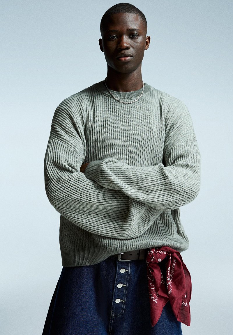 Hellgrüner, gerippter Pullover mit übergroßen Ärmeln, getragen über einem Jeansrock mit großen Knöpfen und einem rot gemusterten Bandana in der Taille.