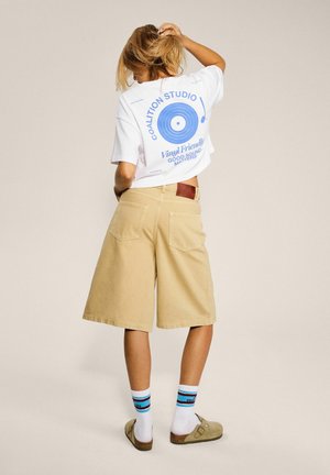 Osoba ubrana w biały krótki t-shirt z niebieskim nadrukiem winylowej płyty, beżowe krótkie spodenki do kolan, paskowane skarpetki i beżowe sandały, stojąca tyłem.