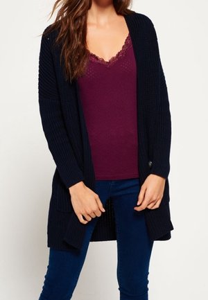 Kvinde iført en mørklilla camisole med blonder, marineblå åben front strikket cardigan og blå jeans, stående med hænderne foldet.