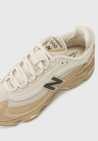 Scarpa sportiva beige realizzata in materiali in rete e suede, con un logo "N" nero, lacci color crema e una suola spessa con dettagli in rilievo.