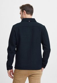 Black collared jacket made from textured fabric, featuring buttoned cuffs and a small tricolor logo at the back neckline.