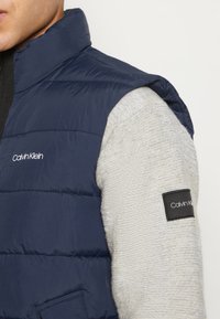 Marineblaue Steppweste mit hohem Kragen, kombiniert mit strukturierten hellgrauen Ärmeln. Mit Calvin Klein-Logo auf der Vorderseite und Ärmelflicken.