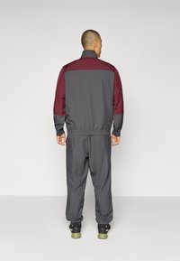 Completo sportivo grigio con accenti marroni, composto da giacca con zip e polsini elasticizzati. I pantaloni della tuta hanno una vita elasticizzata, offrendo una vestibilità comoda.