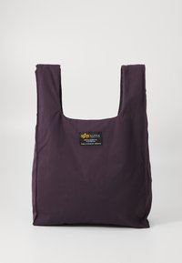 Sac fourre-tout en nylon violet avec une ouverture carrée, arborant une étiquette noire avec logo et texte. Texture lisse, design minimaliste, poignées renforcées.