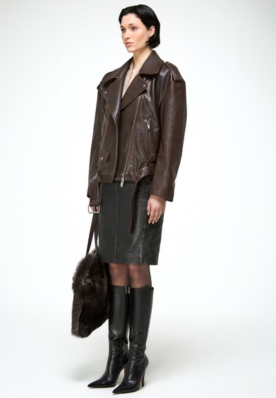 Nő sötétbarna bőrből készült motoros dzsekiben, fekete bőrszoknyában, fekete térdig érő csizmában, és sötét szőrme táskát tartva.