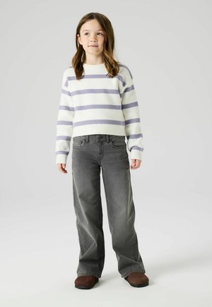 Young girl standing, wearing a white and purple striped sweater, gray wide-leg jeans, and brown slip-on shoes, on a plain light background.