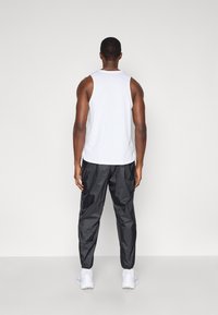 Άνδρας που στέκεται με το πρόσωπο στη σοφίτα, φορώντας λευκό χωρίς μανίκια τοπ, μαύρο αθλητικό παντελόνι, λευκές κάλτσες και λευκά αθλητικά παπούτσια σε απλό φόντο.