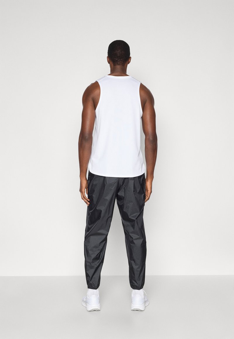 Man staat tegenover een muur en draagt een witte mouwloze top, zwarte sportbroek, witte sokken en witte sneakers op een effen achtergrond.