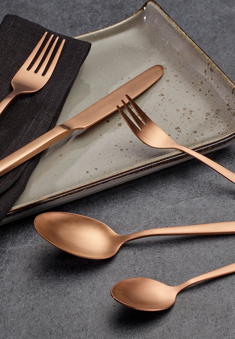 Das kupferfarbene Besteckset umfasst ein Messer, zwei Gabeln und zwei Löffel mit glatter Oberfläche, die auf einem gesprenkelten hellgrauen Teller und einer schwarzen Serviette liegen.