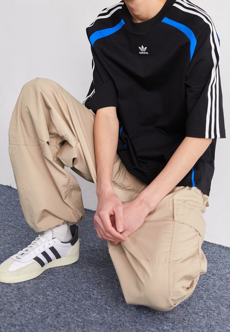 Černé tričko s modrými detaily, bílé pruhy Adidas, béžové cargo kalhoty a bílé tenisky s černými pruhy. Jednoduché pozadí.