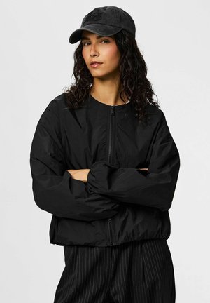 Frau mit lockigem Haar, die eine schwarze Jacke mit Reißverschluss, schwarze Nadelstreifenhose und eine schwarze Baseballkappe trägt, mit verschränkten Armen und neutralem Gesichtsausdruck.