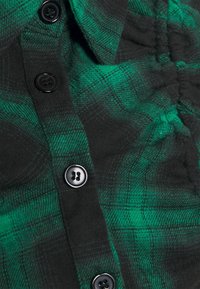 Camicia a bottoni caratterizzata da un motivo a quadri verde e nero. Realizzata in tessuto morbido, con bottoni neri rotondi e dettagli in tessuto arricciato.