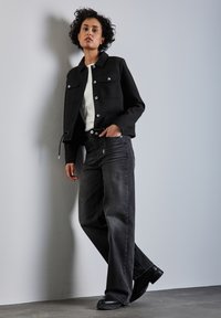 Veste noire courte avec des boutons argentés, t-shirt blanc et jeans larges gris foncé, associés à des bottines noires. Tissu texturé, look décontracté.