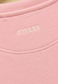 Gros plan sur un tissu rose avec un col côtelé et le texte brodé blanc "JOULES" centré sous la couture de l'encolure.