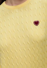 Pull jaune en maille torsadée avec un petit patch cœur rouge sur la poitrine gauche, doté d'un col rond côtelé.