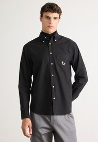 Hombre con camisa negra de botones sobre un suéter de cuello alto negro y pantalones grises, de pie con una mano detrás de la espalda sobre un fondo blanco.