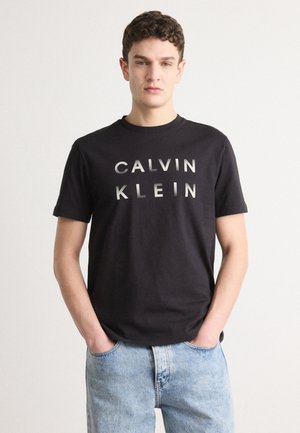 Black cotton t-shirt with "CALVIN KLEIN" printed in bold silver letters on the front. Model wears light blue denim jeans.