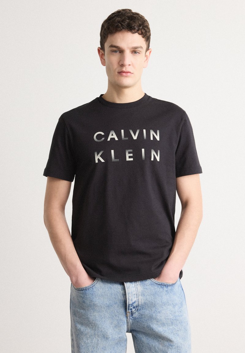 Tricou din bumbac negru cu "CALVIN KLEIN" tipărit cu litere argintii îngroșate pe față. Modelul poartă blugi din denim albastru deschis.