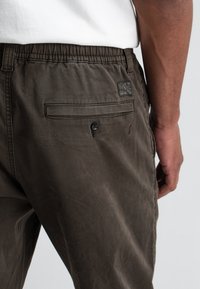 Donkergroene olijfkleurige broek van zacht stof met een elastische tailleband, voorzien van een achterzak, knoopdetail en een tekstuurpatch-logo.