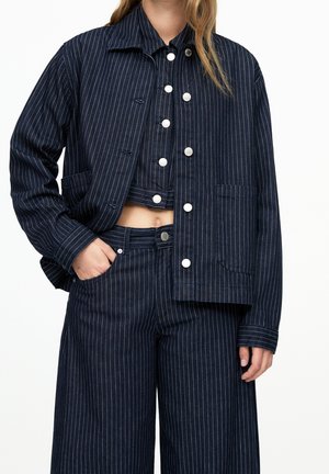 Femme portant une veste bleu marine à fines rayures, un gilet court boutonné et un pantalon large, main dans la poche, sur fond blanc.